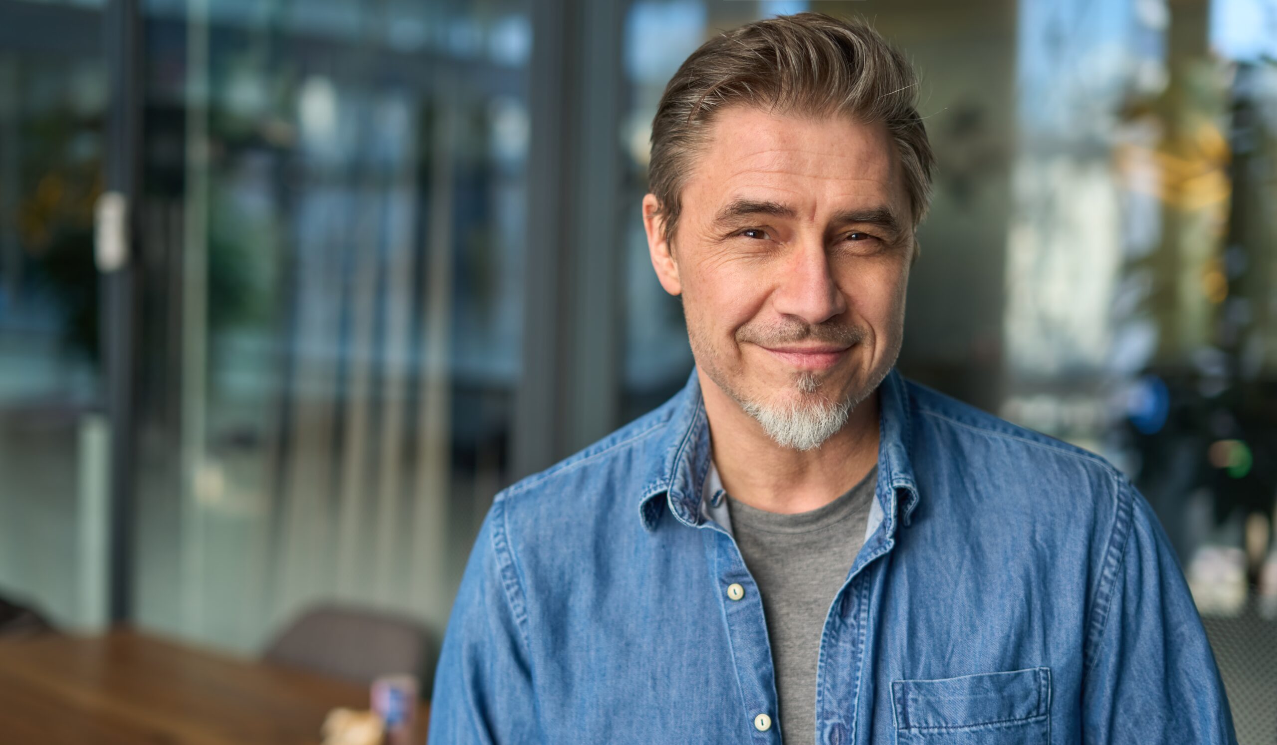Portrait of happy middle aged man in glasses, standing in office lobby wearing casual shirt.